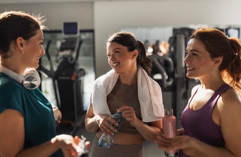 Three cheerful young women resting in gym and talking after good workout, holding bottles with water and protein bars. Sport and healthy lifestyle concept