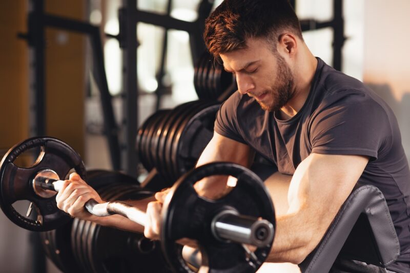 Handsome man doing biceps lifting barbell on bench in a gym